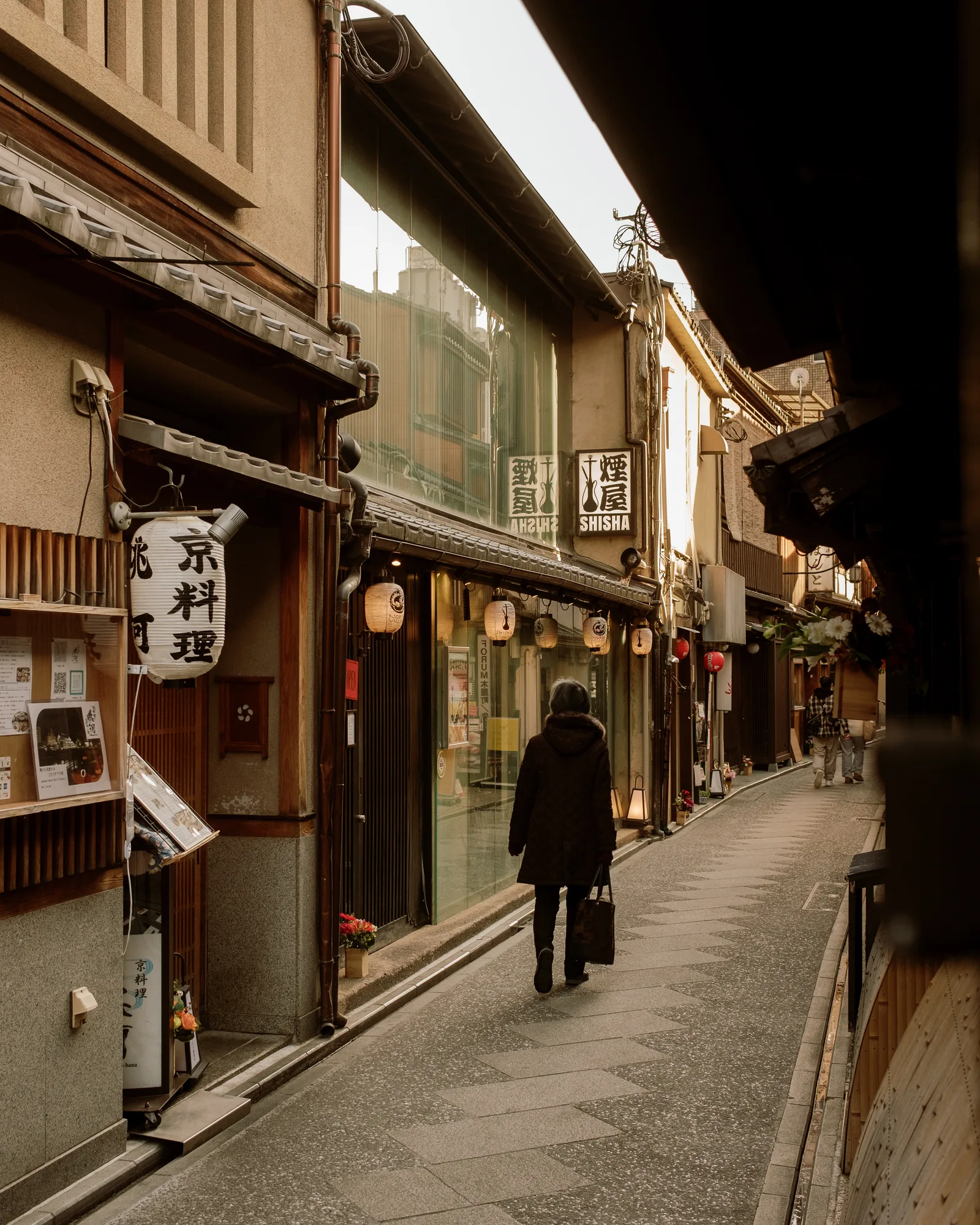 Woman in dark clothes walking down a narrow Kyoto lane past wooden shopfronts