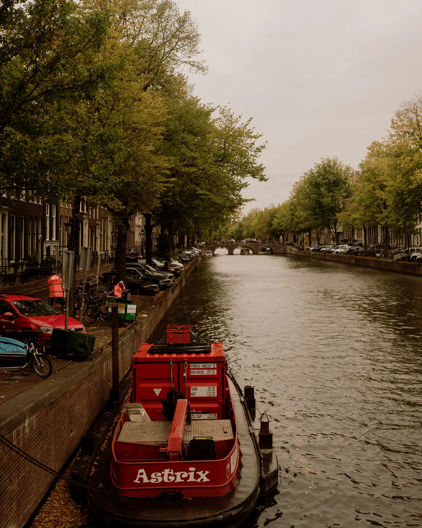 Amsterdam canal with a red work barge moored beside a tree-lined quay under a gray sky