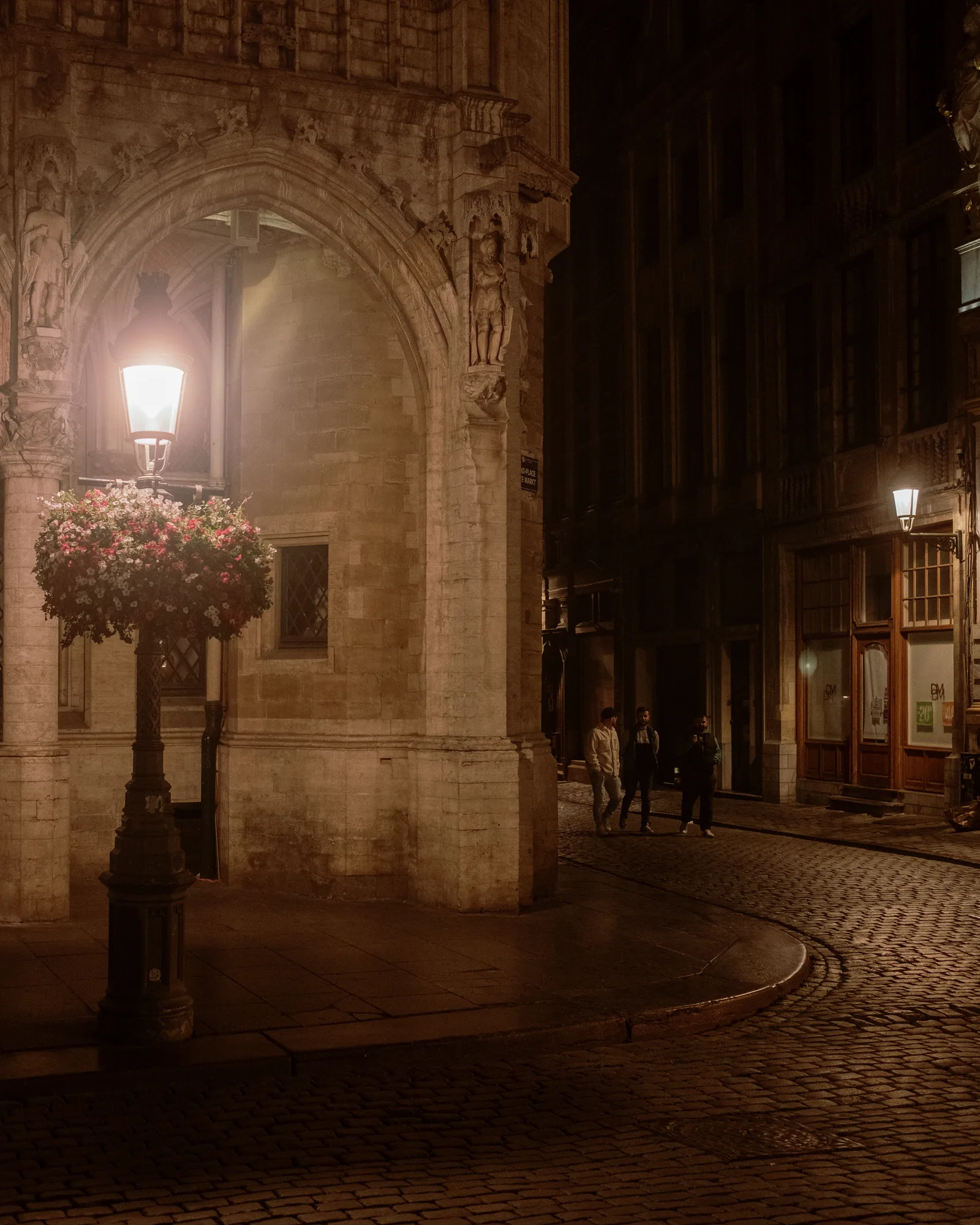 Lamp post with flower baskets beside a carved stone arch on a cobbled Brussels street at night