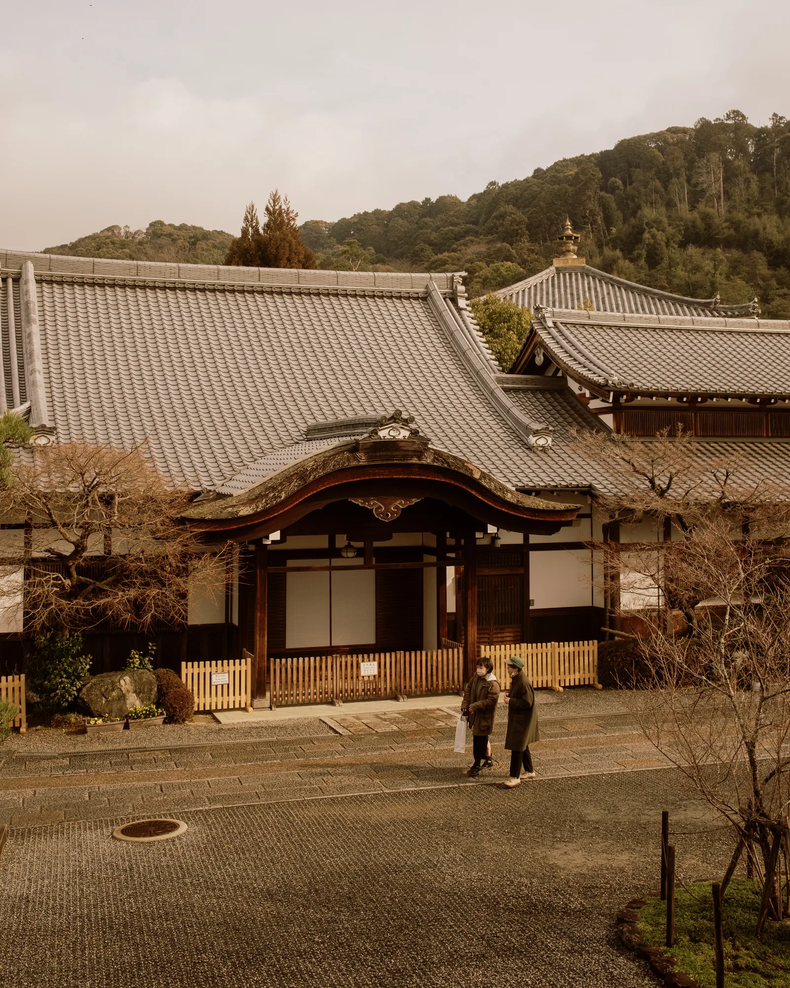 Traditional Kyoto temple building with a tiled roof and two people walking past, wooded hill behind