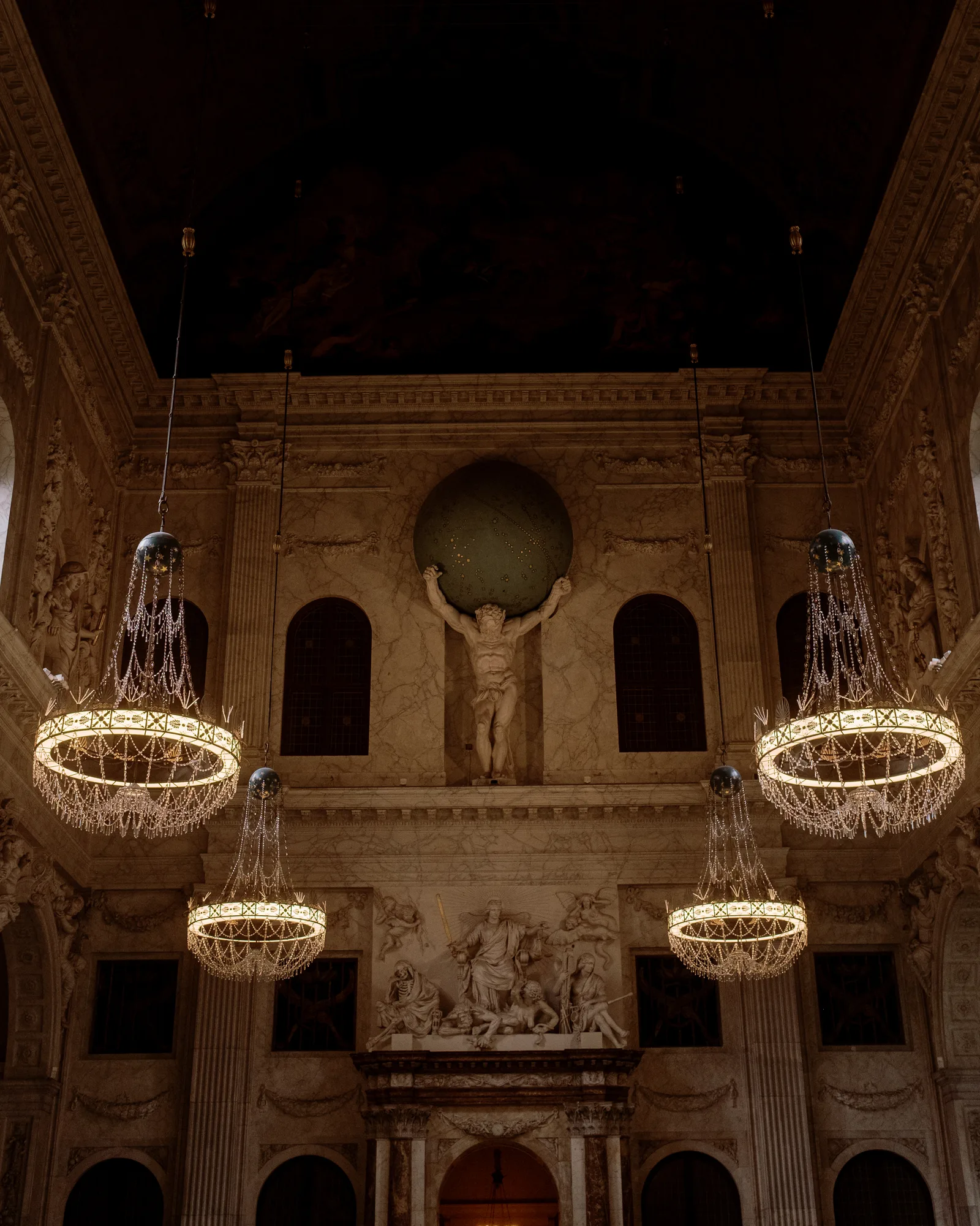 Grand stone hall in Amsterdam with a sculpted figure above the wall and glowing crystal chandeliers