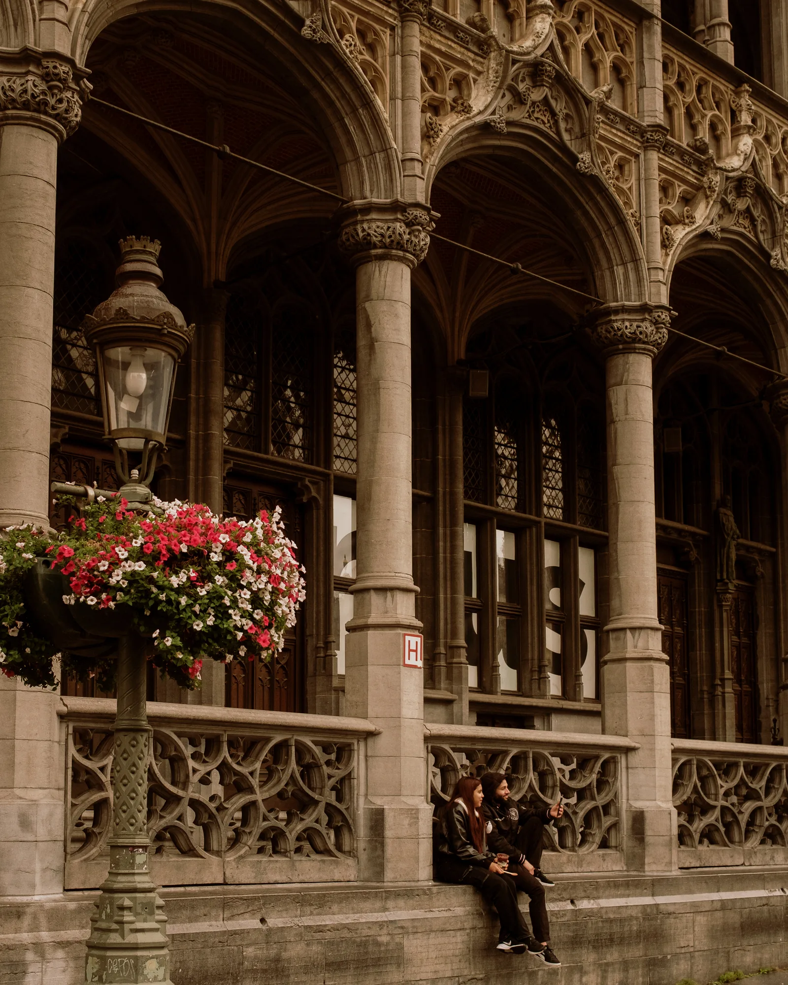 Two people sitting on a stone ledge beneath ornate Gothic arcades and a lamp post with flowers, Brussels