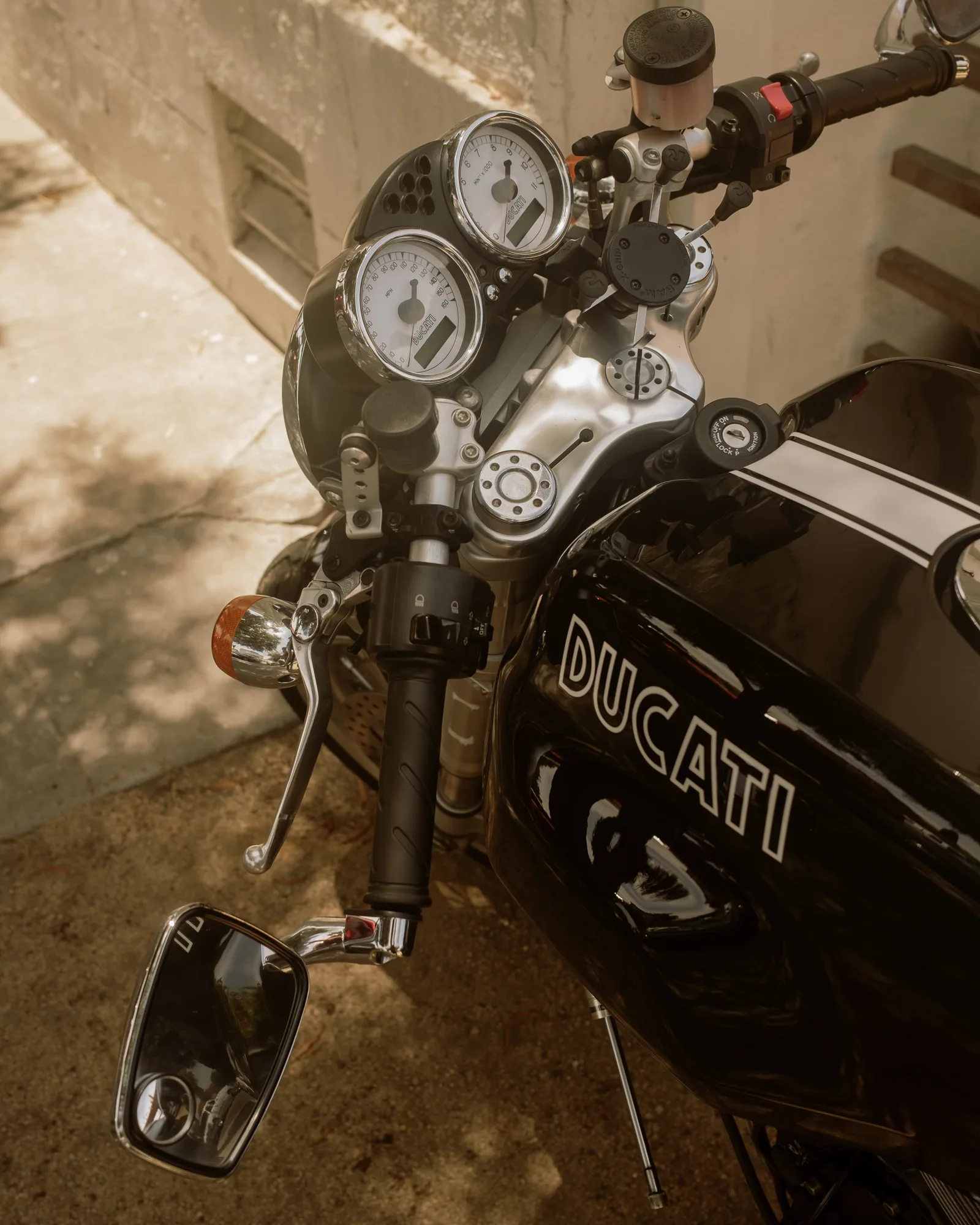Black Ducati motorcycle gauges and handlebars in dappled light against a concrete wall, Barrio Logan