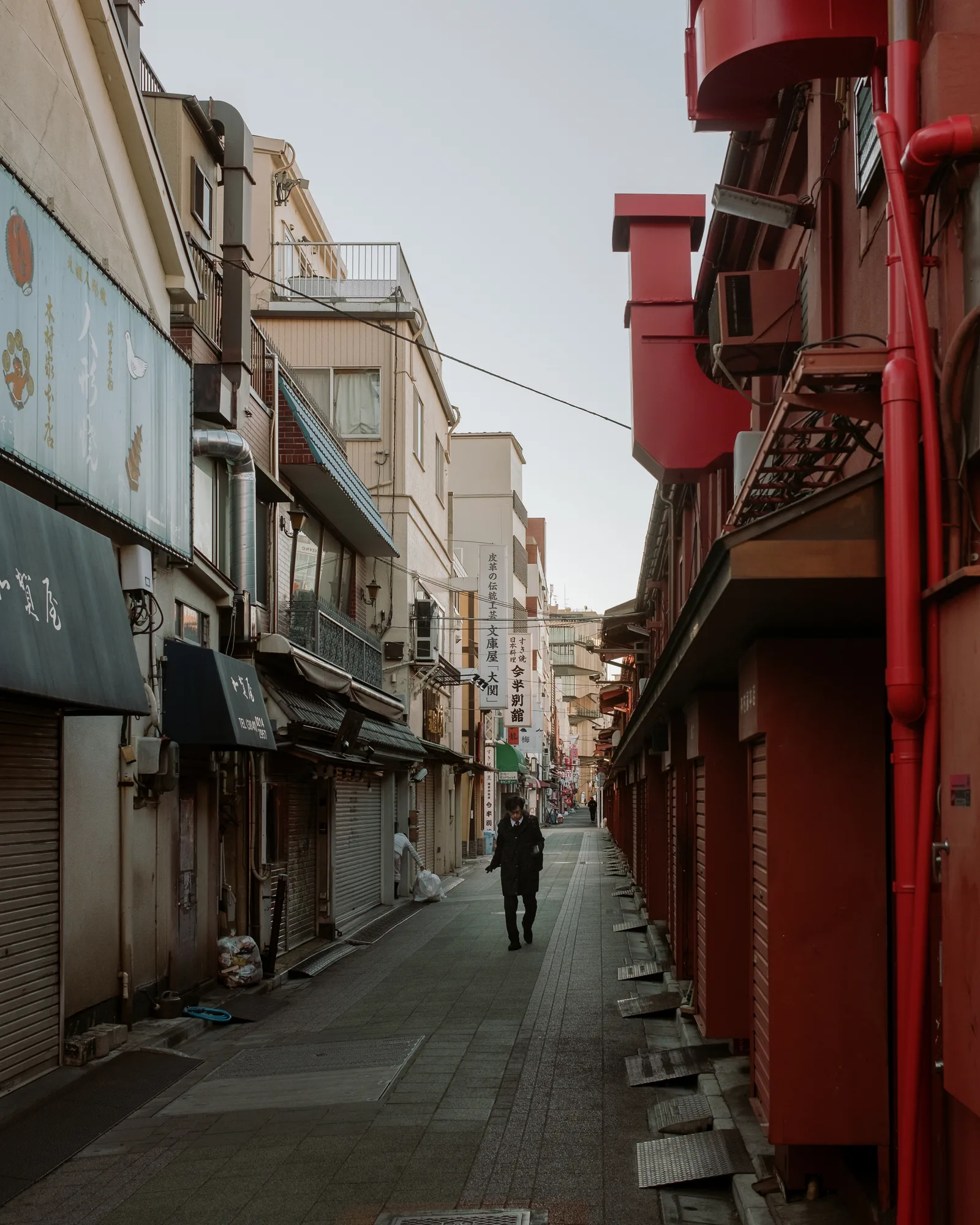 Person walking down a narrow Asakusa lane between shuttered shops and red-painted walls, Tokyo