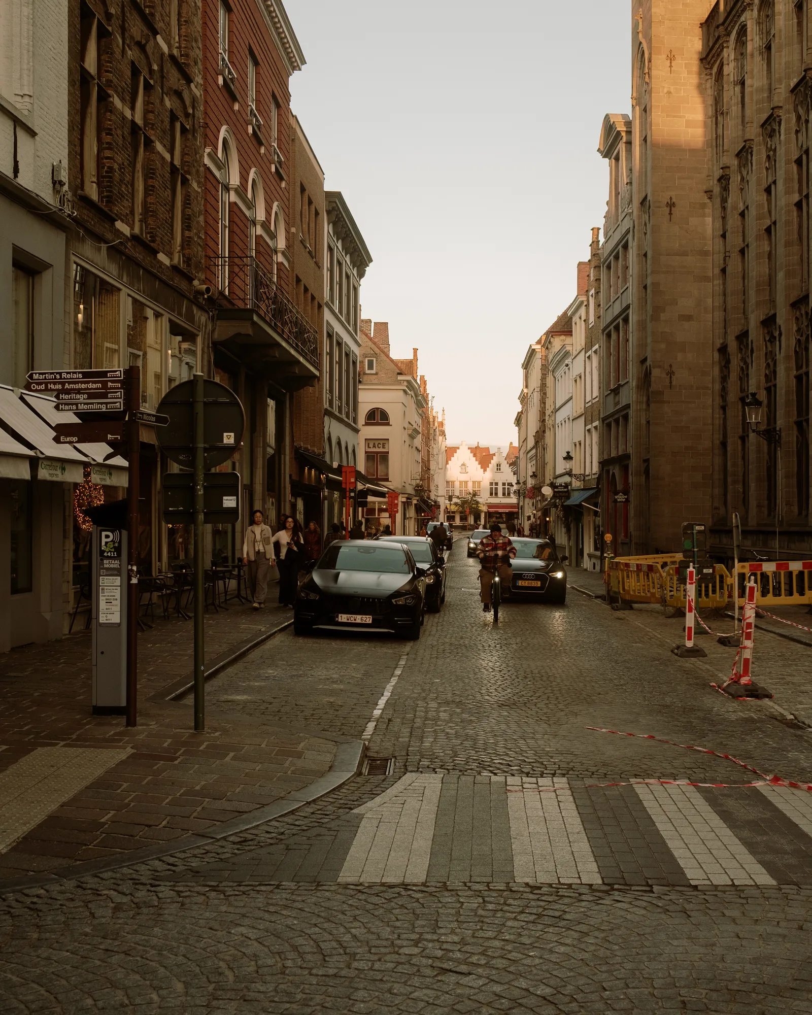Cobbled Bruges street with a crosswalk, cars and shopfronts in warm evening light
