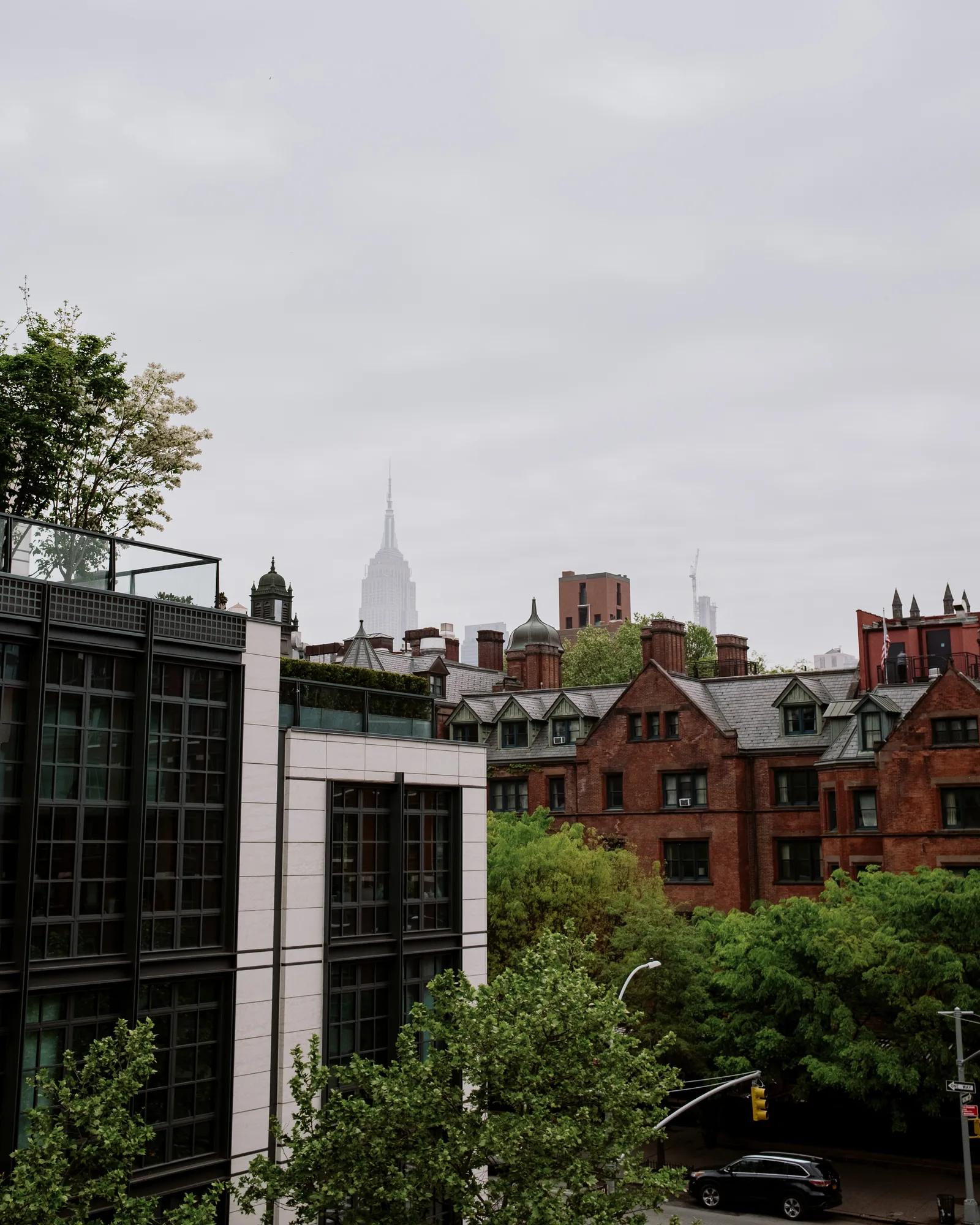 Empire State Building tip above red-brick rooftops and green trees on an overcast day, New York