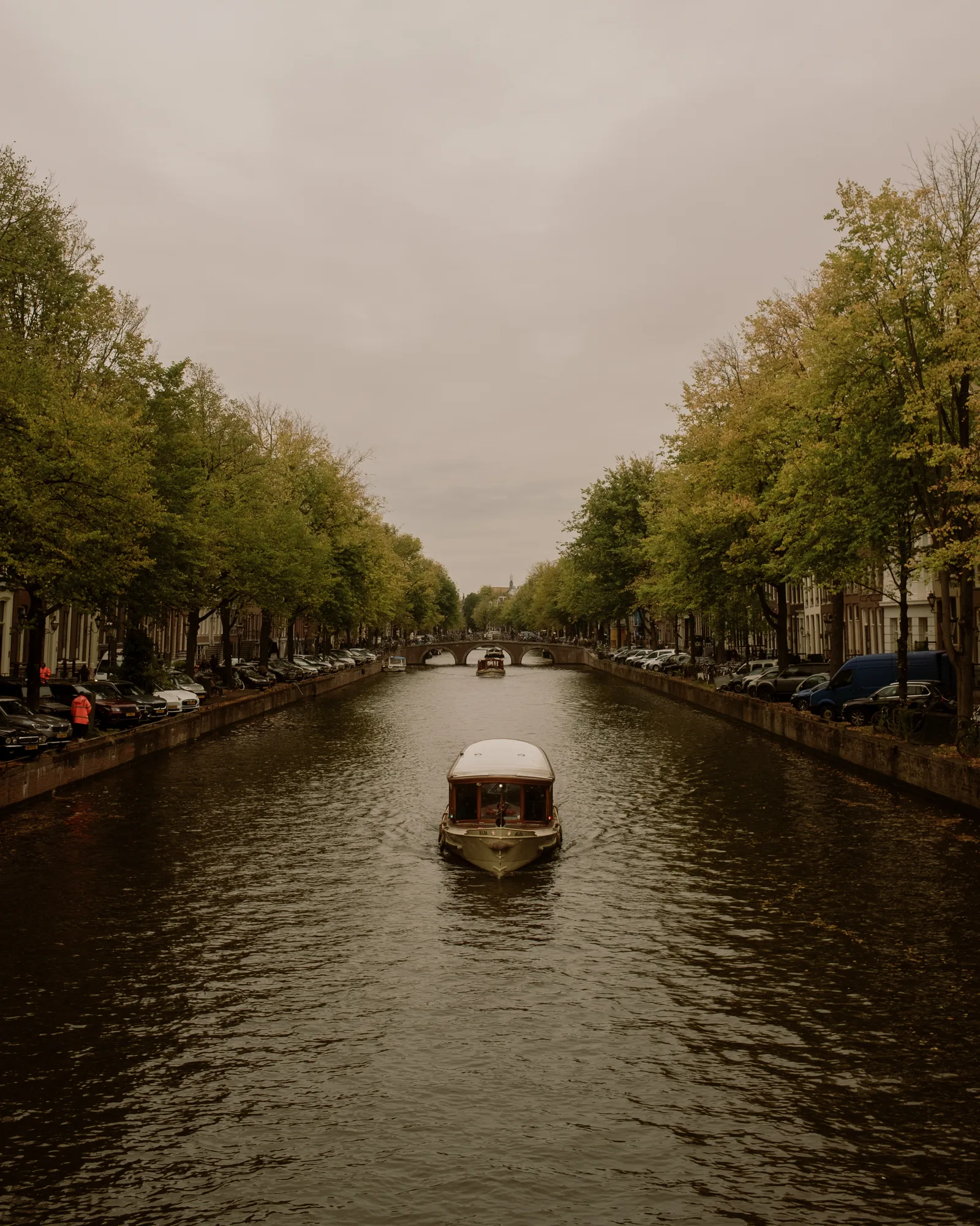 Amsterdam canal on an overcast day, a small boat heading toward a distant bridge between tree-lined quays