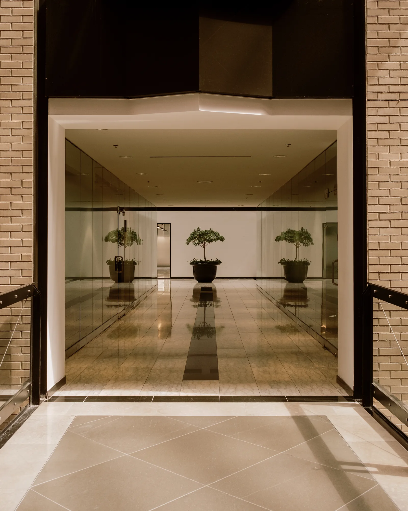 Glass-walled lobby with potted trees and a polished floor seen through a brick opening, Montreal