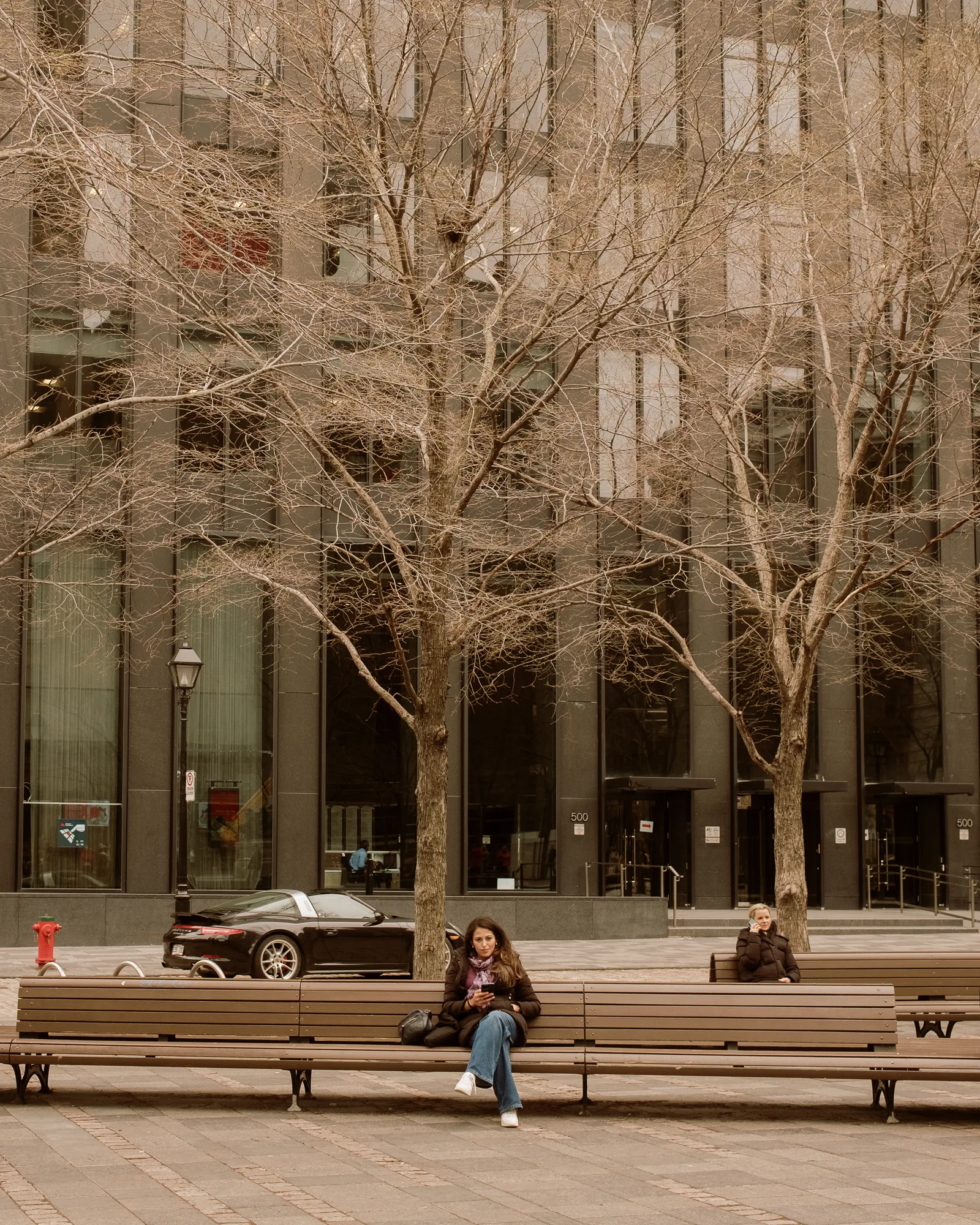 Person sitting on a bench in a plaza of bare trees and glass towers, Montreal