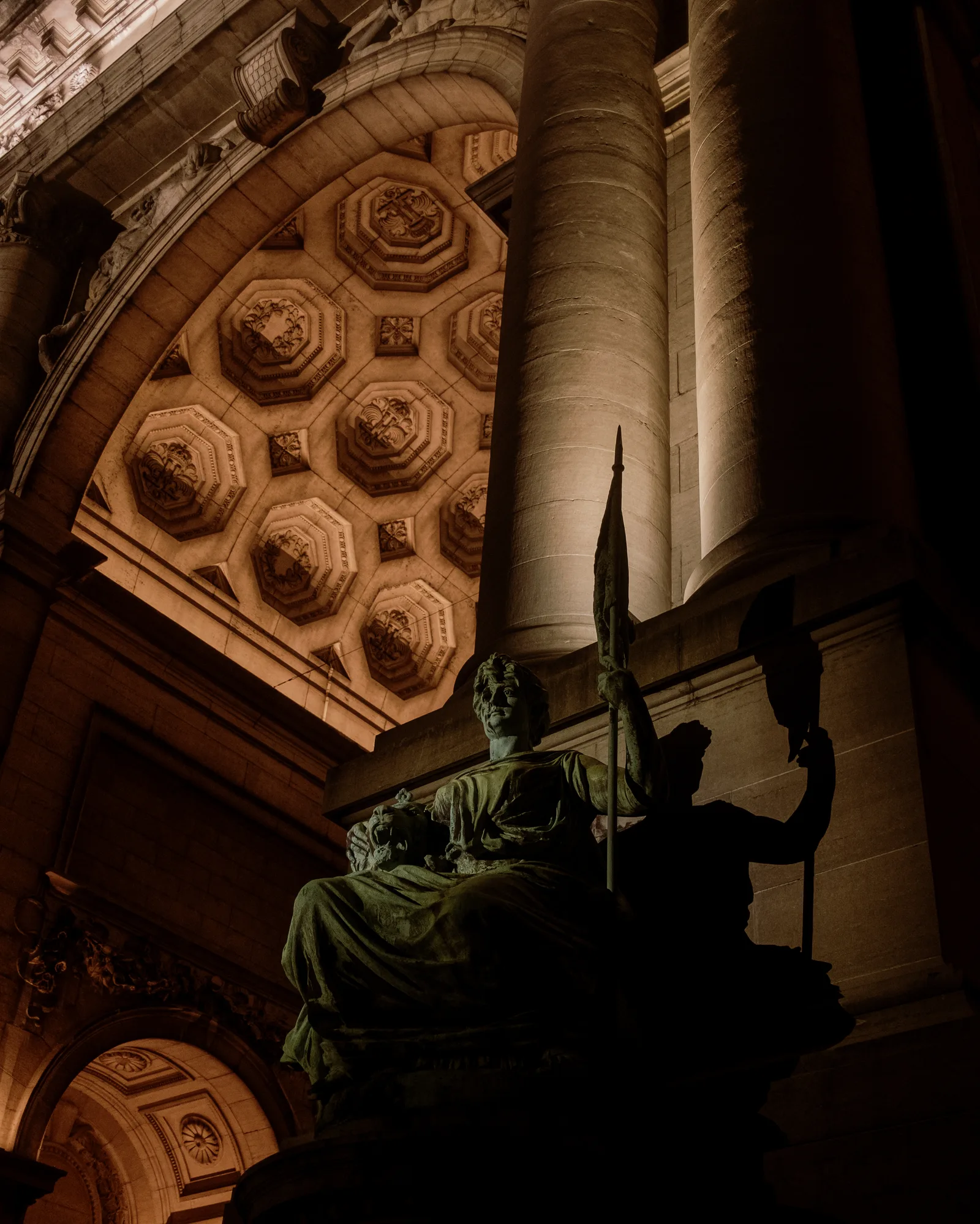 Statue in silhouette beside a coffered stone archway lit from below, Brussels