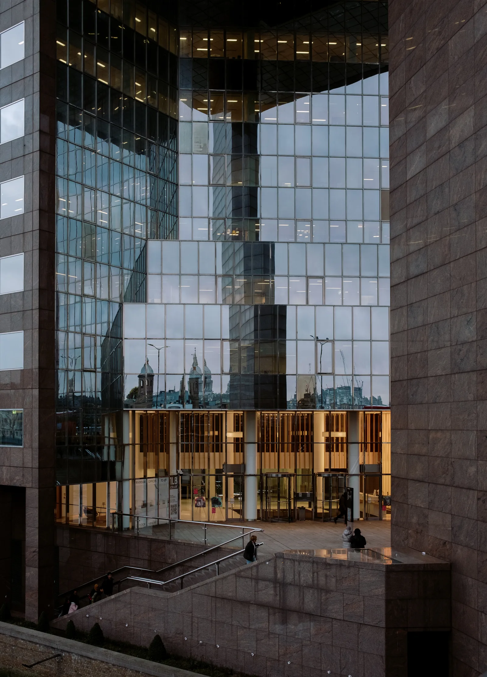 Glass office building reflecting the sky above a lit ground-floor lobby, London