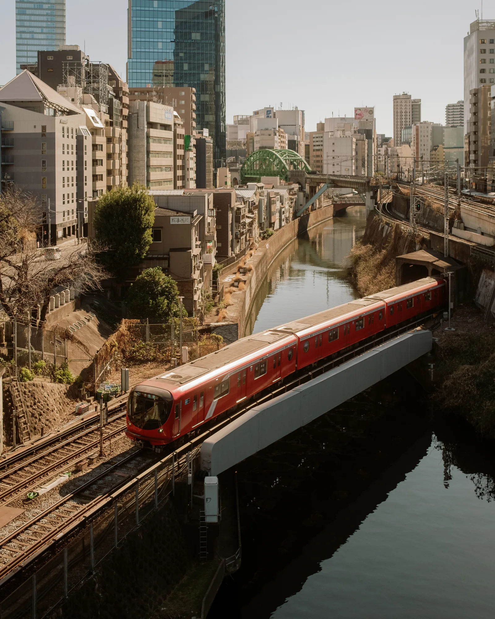 Red train passing along a canal in Tokyo at golden hour, high-rises behind