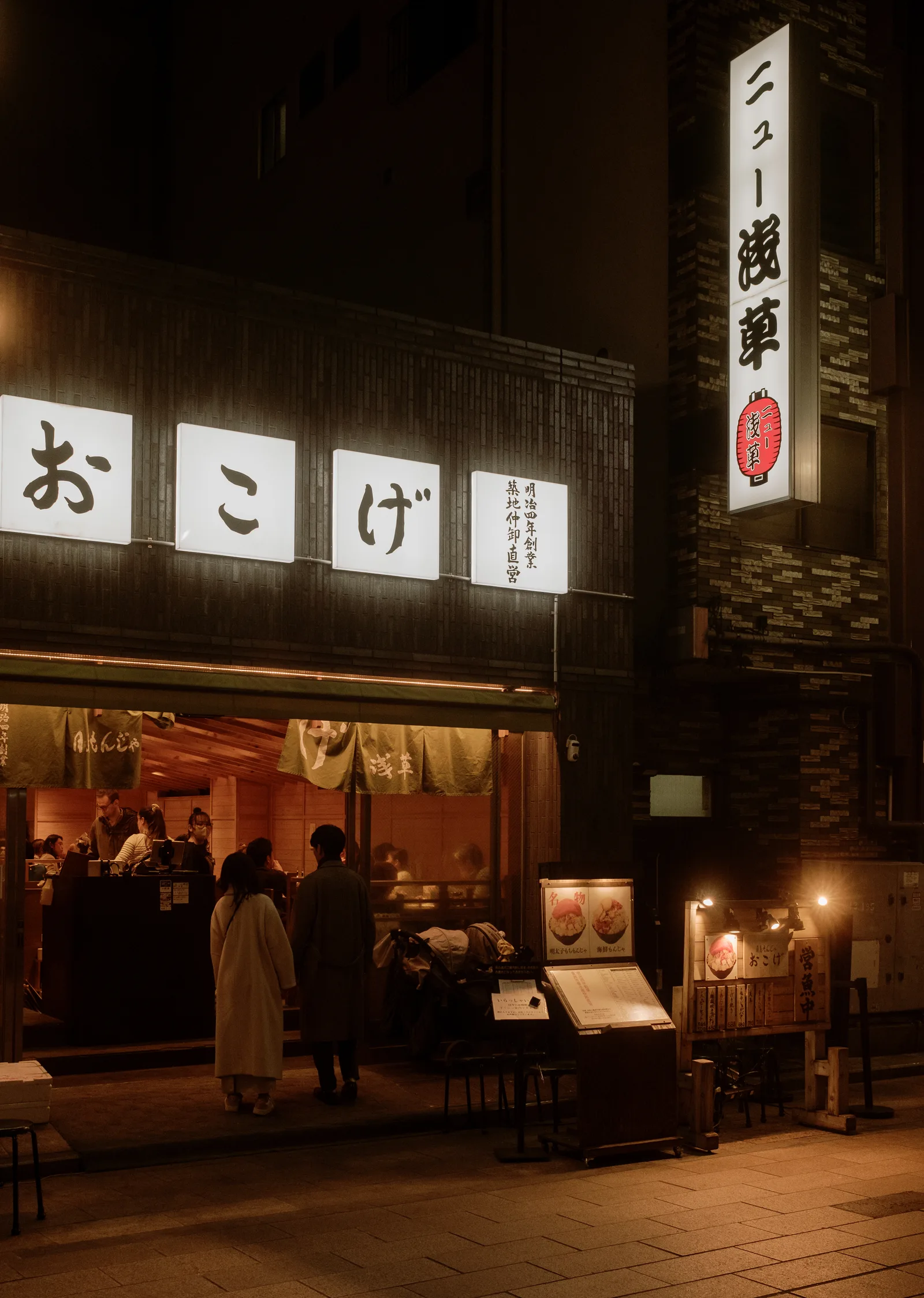 Lit restaurant entrance with white kana signs and diners inside, Asakusa, Tokyo at night