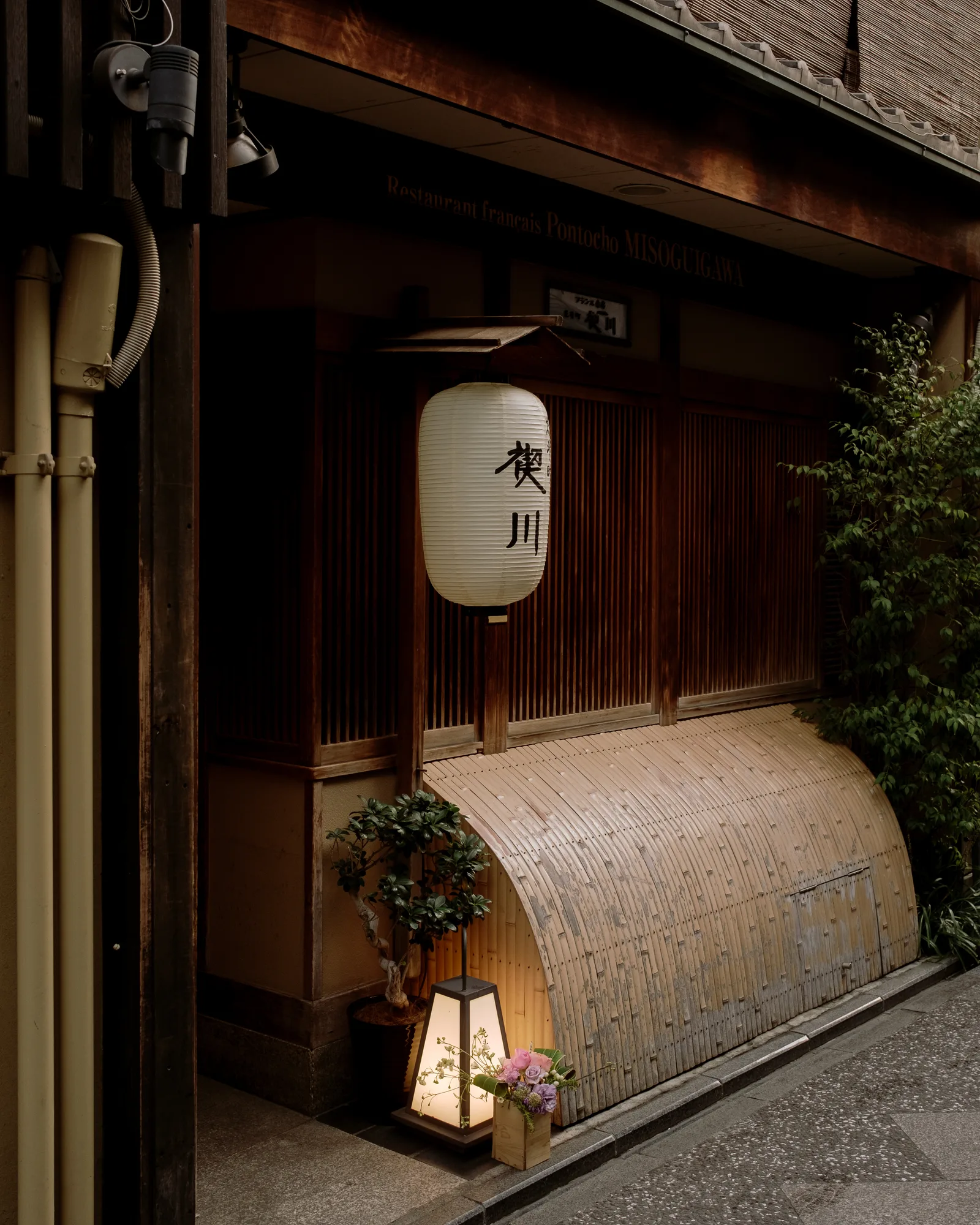 Kyoto restaurant entrance with a round lantern, a curved bamboo screen and a glowing floor lamp