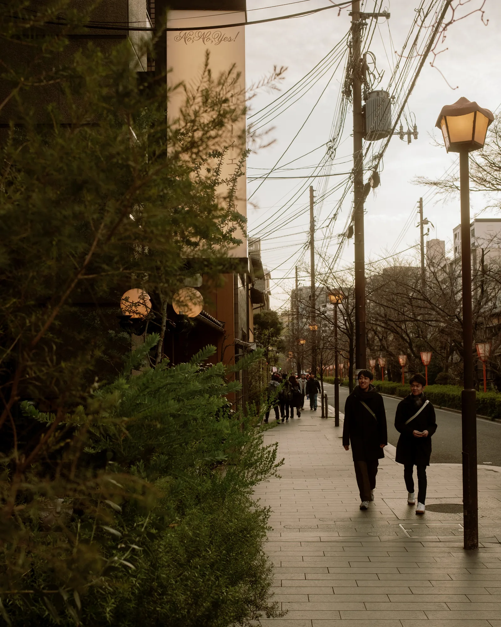 Two people walking down a Kyoto sidewalk lined with lamps and overhead wires at dusk