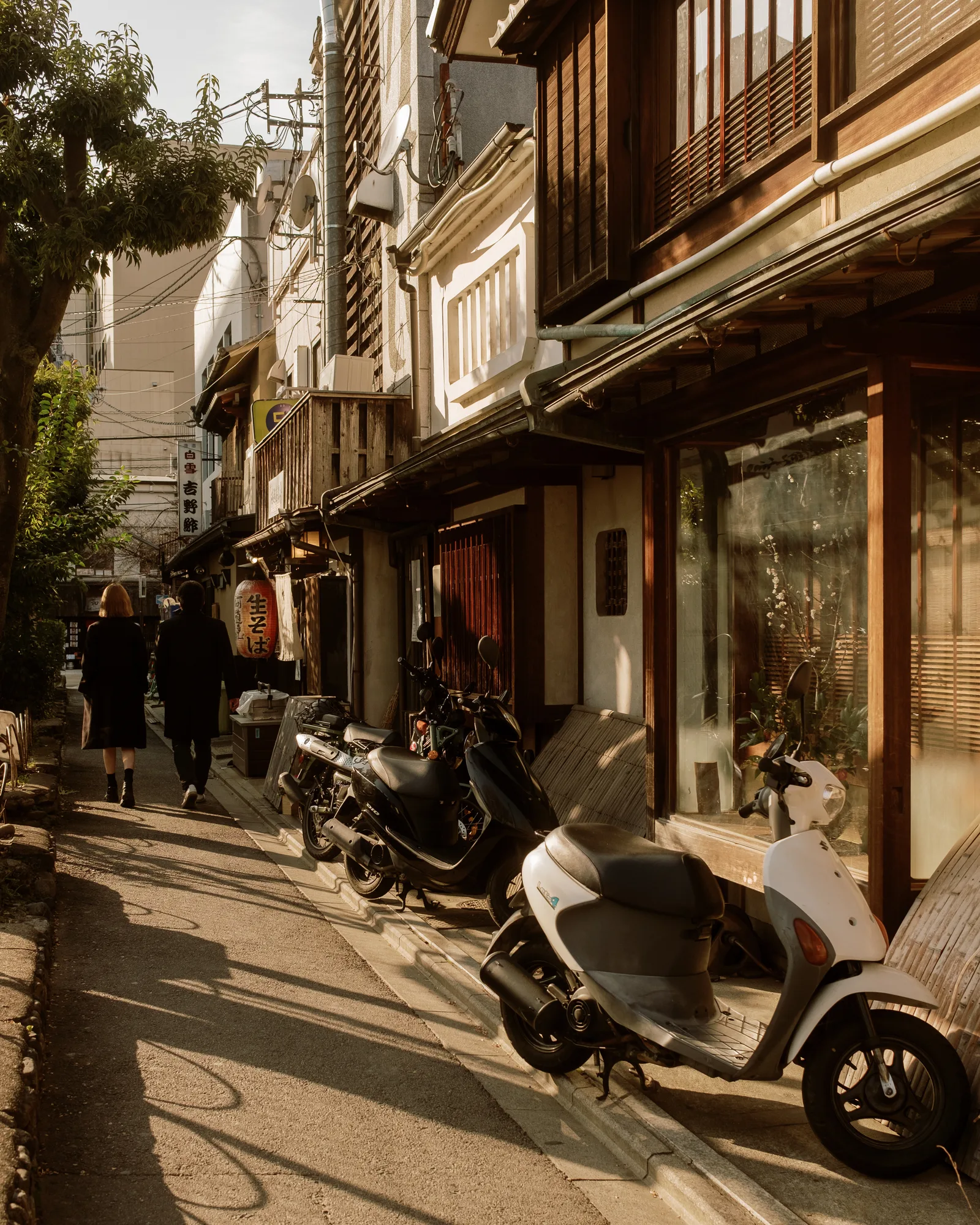 Sunlit Kyoto lane with parked scooters and two people walking past wooden shopfronts
