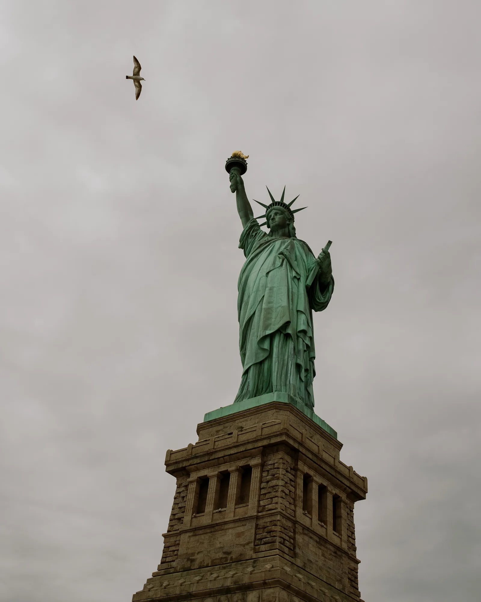 Statue of Liberty above its pedestal, a bird in flight in a gray sky
