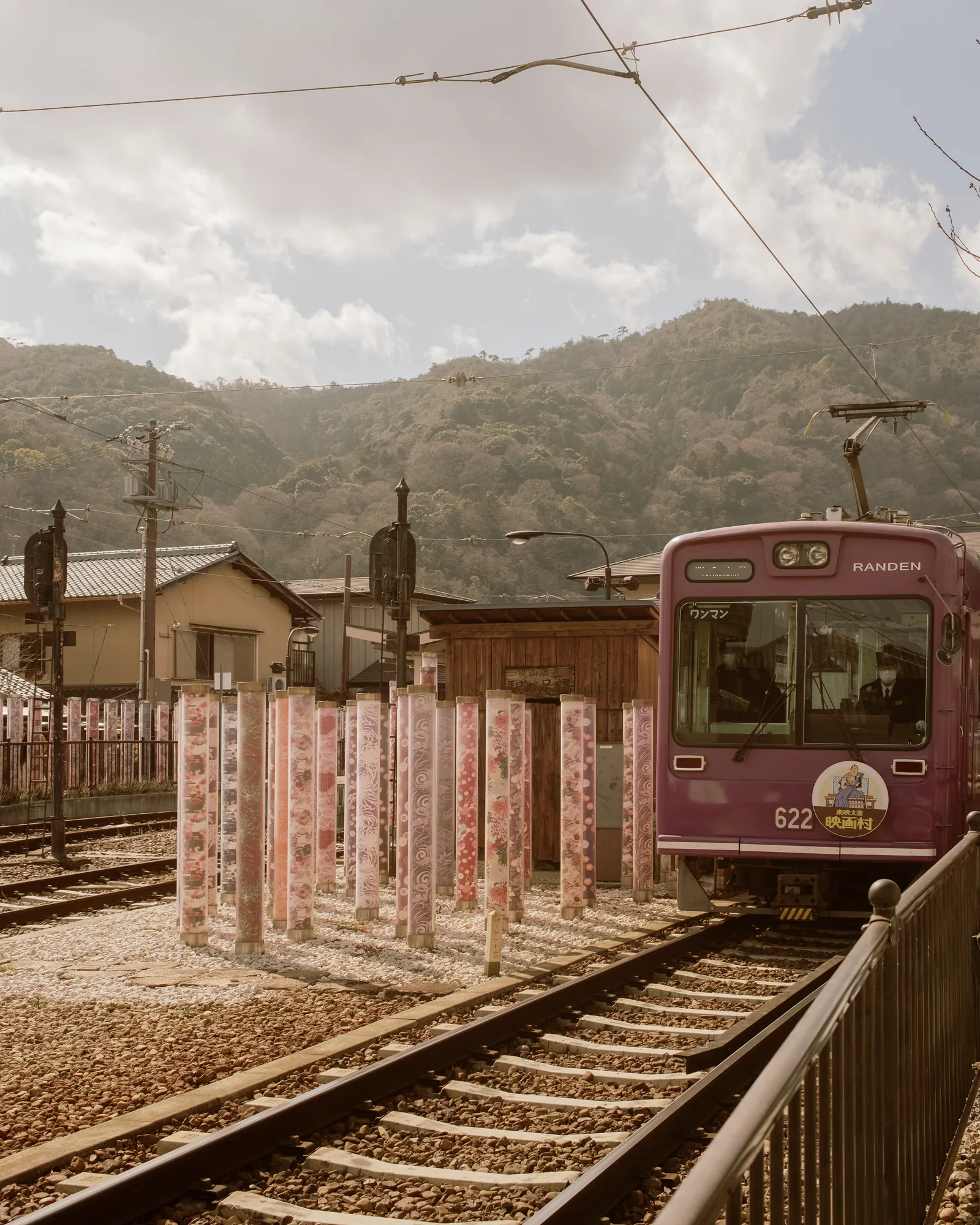 Purple tram at a station beside a display of pink-and-white banners, hills behind, Kyoto