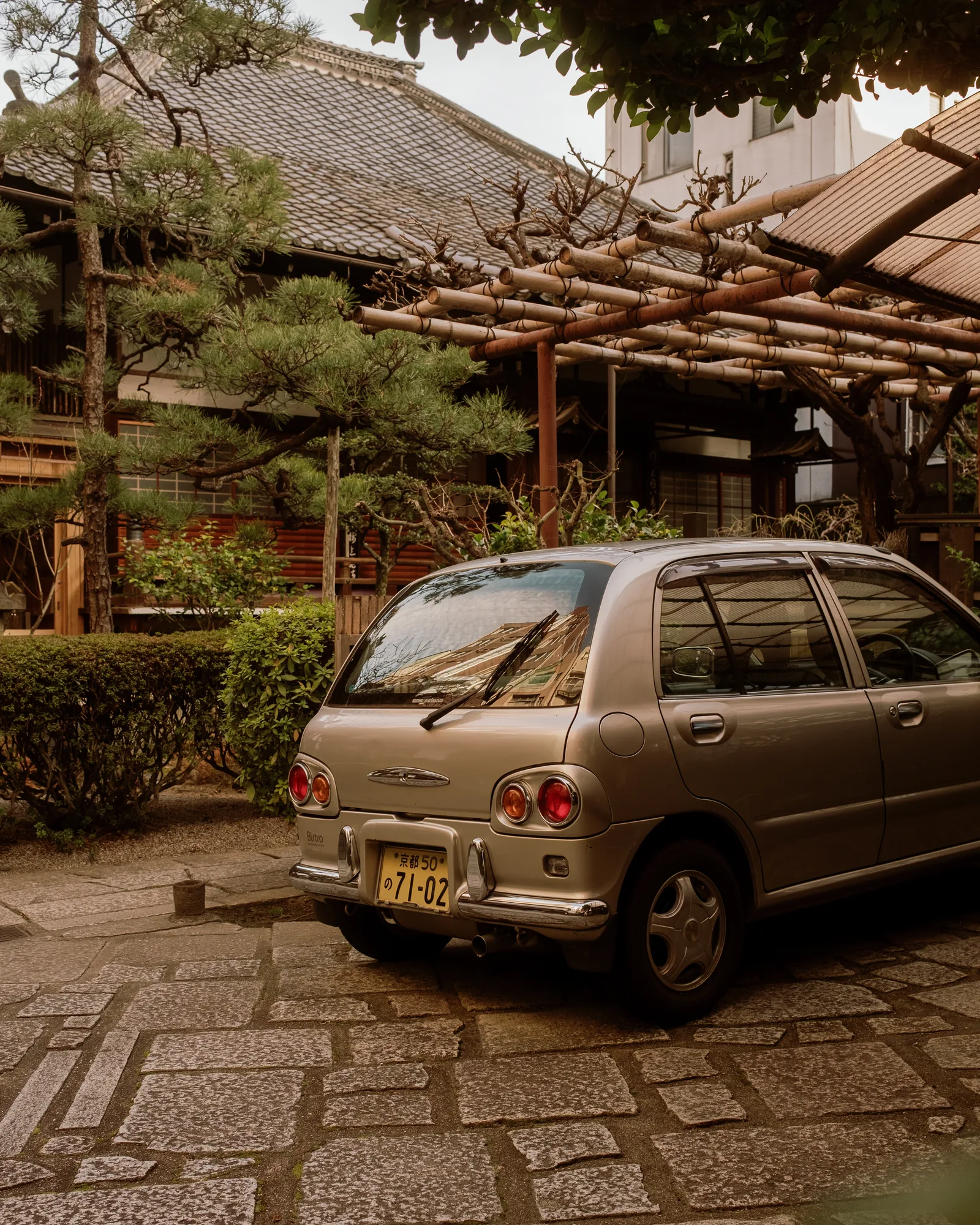 Beige hatchback parked on stone paving beneath a pine and wooden trellis, Kyoto