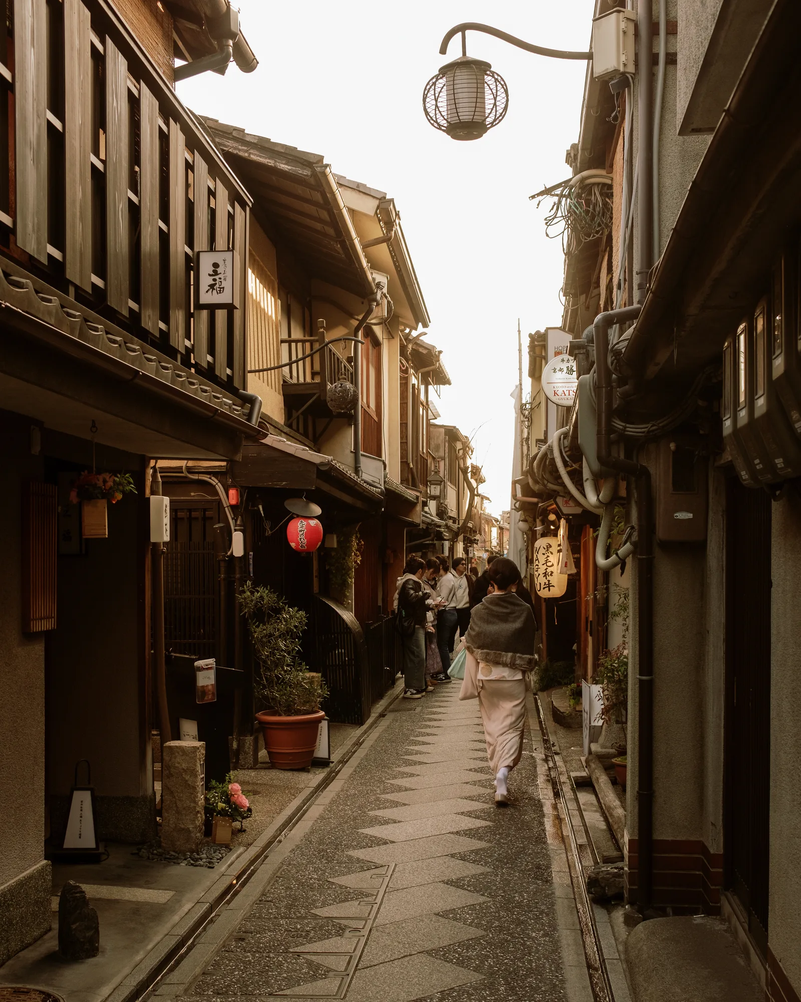 Person walking away down a narrow Kyoto alley of wooden shopfronts, lanterns overhead