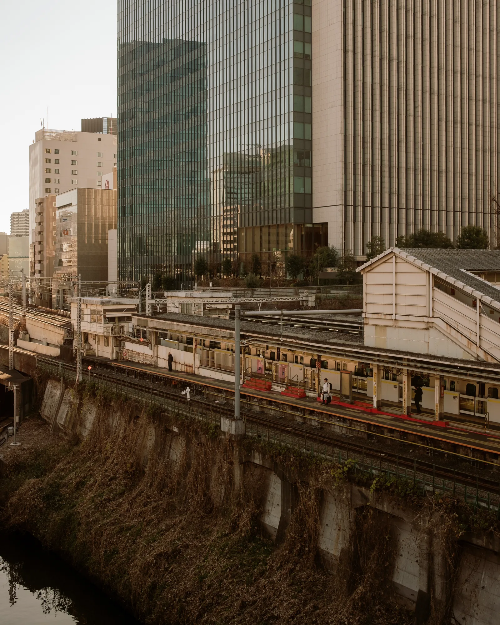 Commuter train at a station platform beside a canal, glass towers behind, Tokyo
