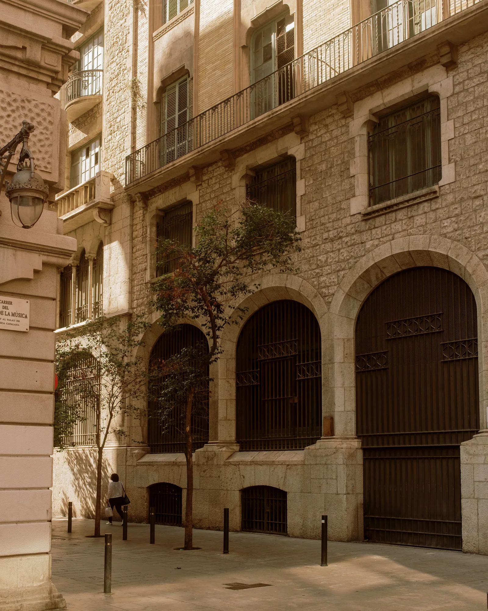 Stone building with tall arched black gates, bollards and a person walking in low sun, Barcelona