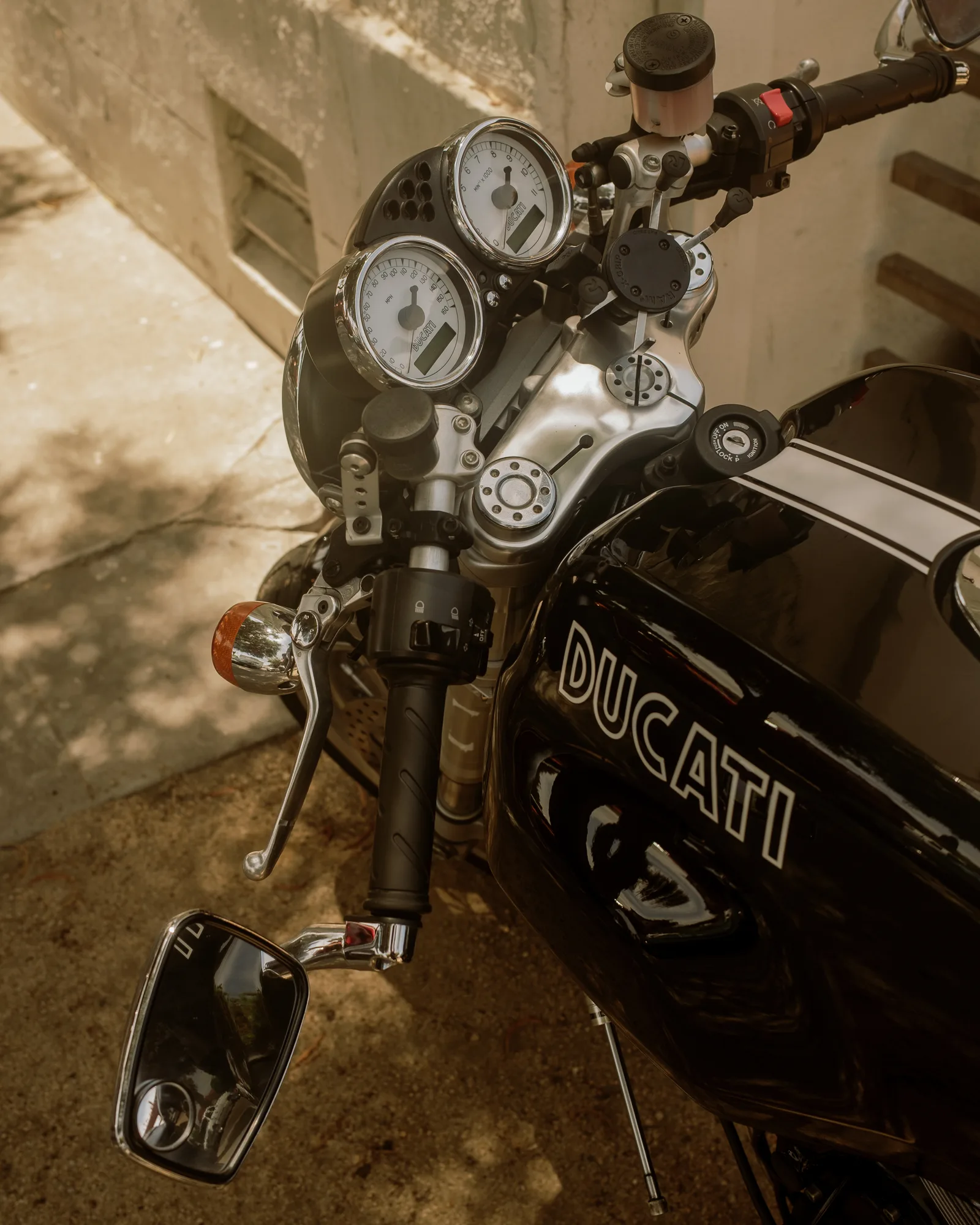 Black Ducati motorcycle gauges and handlebars in dappled light against a concrete wall, Barrio Logan